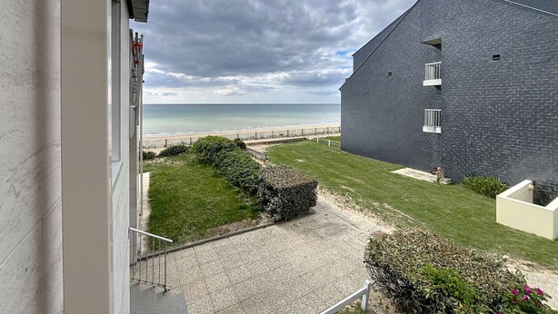 A view from a balcony overlooking a beach, a grassy area, and a modern building with a dark facade. - Charming 1-bed apt, sleeps 4, balcony, parking, wi-fi (Jullouville)