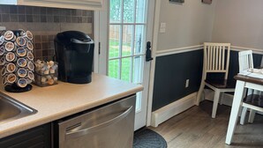 Una cocina compacta con lavavajillas, cafetera y un soporte para cápsulas de café. Hay una zona de comedor con una mesa y sillas, y una puerta que da al exterior.
