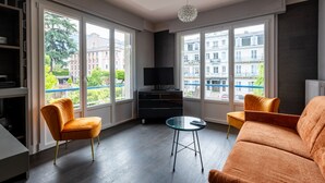 A modern living room with large windows, a flat-screen TV, a glass coffee table, and two orange armchairs.