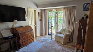Una habitación con un aparador de madera, un escritorio con una lámpara, una silla, un televisor montado en la pared y un ventanal corredizo que da a un espacio al aire libre.