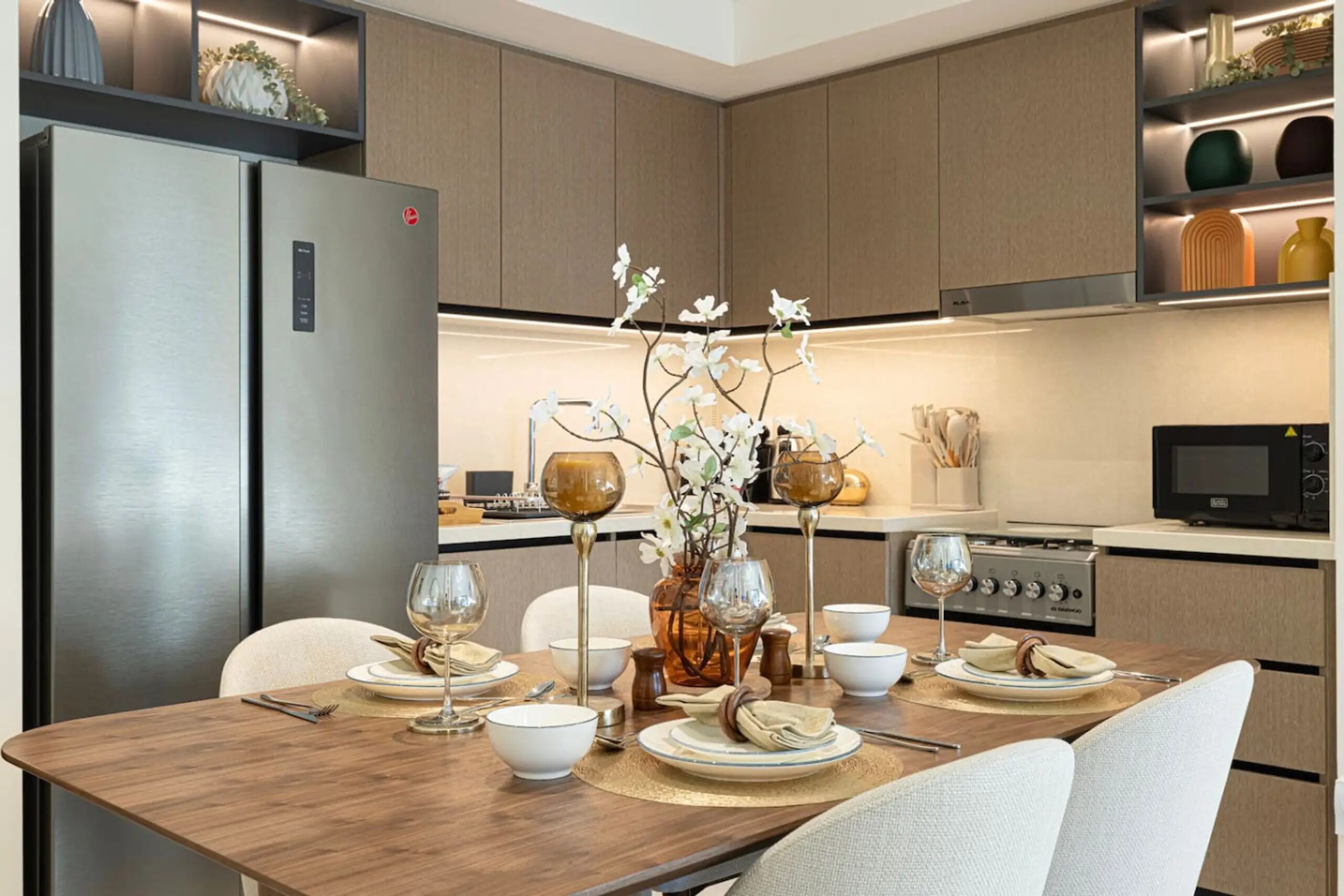 Une cuisine moderne équipée d’appareils électroménagers en acier inoxydable, une table à manger en bois dressée avec de l’argenterie blanche, des bols et des verres à vin, et un vase contenant des fleurs blanches.