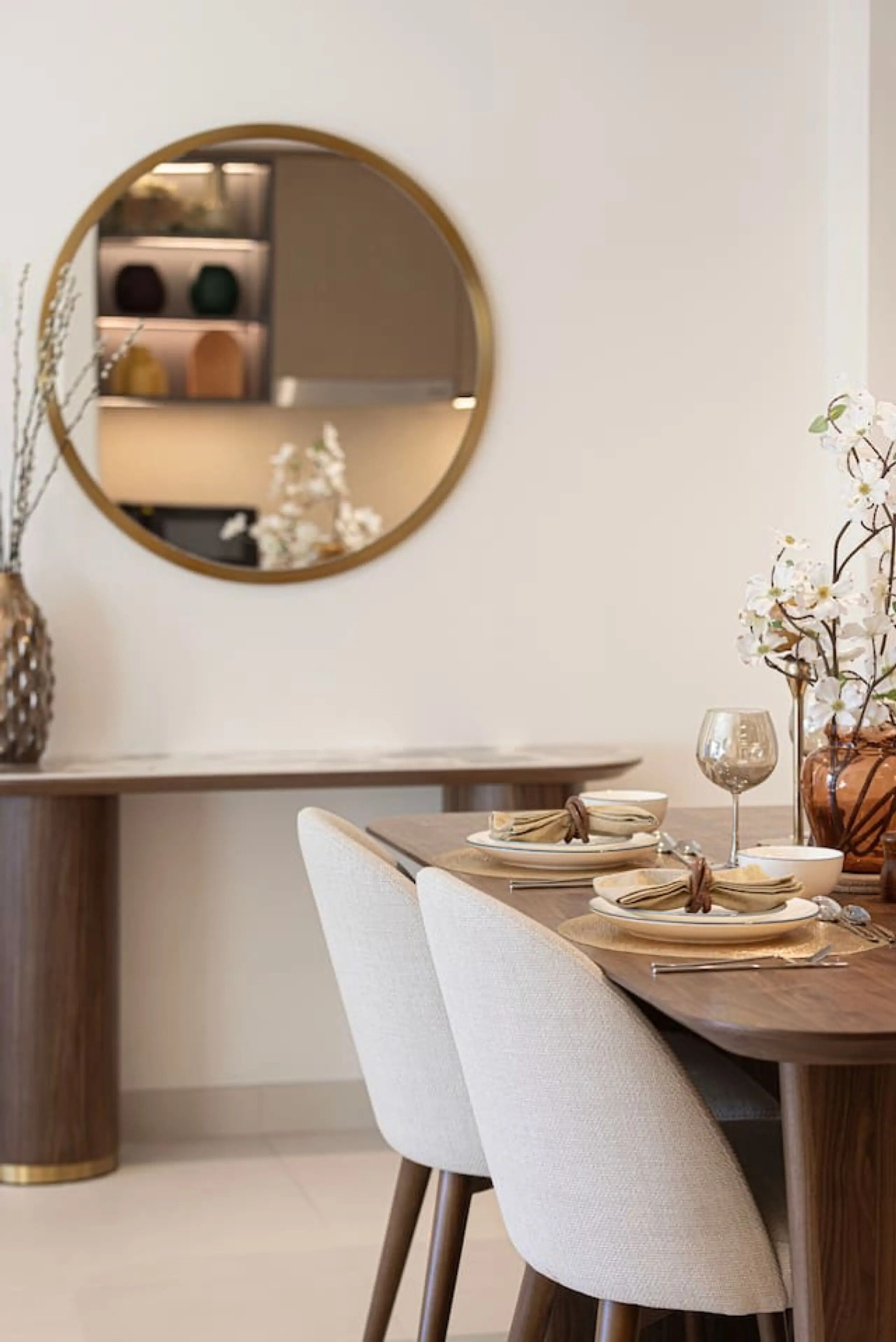 Un coin repas agrémenté d’un miroir rond, d’un vase avec des fleurs blanches et d’une table en bois mise pour deux personnes.