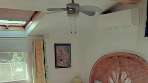 Une chambre à coucher comprenant un lit, un ventilateur de plafond, une fenêtre avec des rideaux, un tableau accroché au mur et une tête de lit en osier.