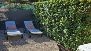 Un espace patio aménagé avec deux chaises longues et une table, entouré de gravier et d’une haie.