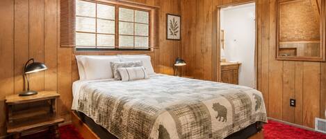 Un dormitorio con paneles de madera, una cama, dos lámparas de noche, una ventana con persianas, un cuadro enmarcado y una puerta con espejo.