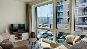 Een moderne woonkamer met een grote televisie, comfortabele zithoek en uitzicht op een jachthaven en gebouwen.