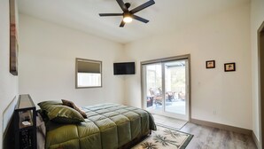 A bedroom with a bed, a ceiling fan, a TV, a window with blinds, a door leading to a balcony, and two framed pictures on the wall.