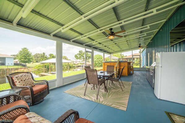A covered patio area with wicker furniture, a dining table, and a kitchen area with a refrigerator.