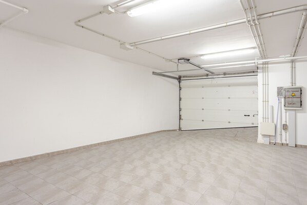 A clean, empty garage with white walls and a tiled floor, featuring overhead fluorescent lighting and visible plumbing.