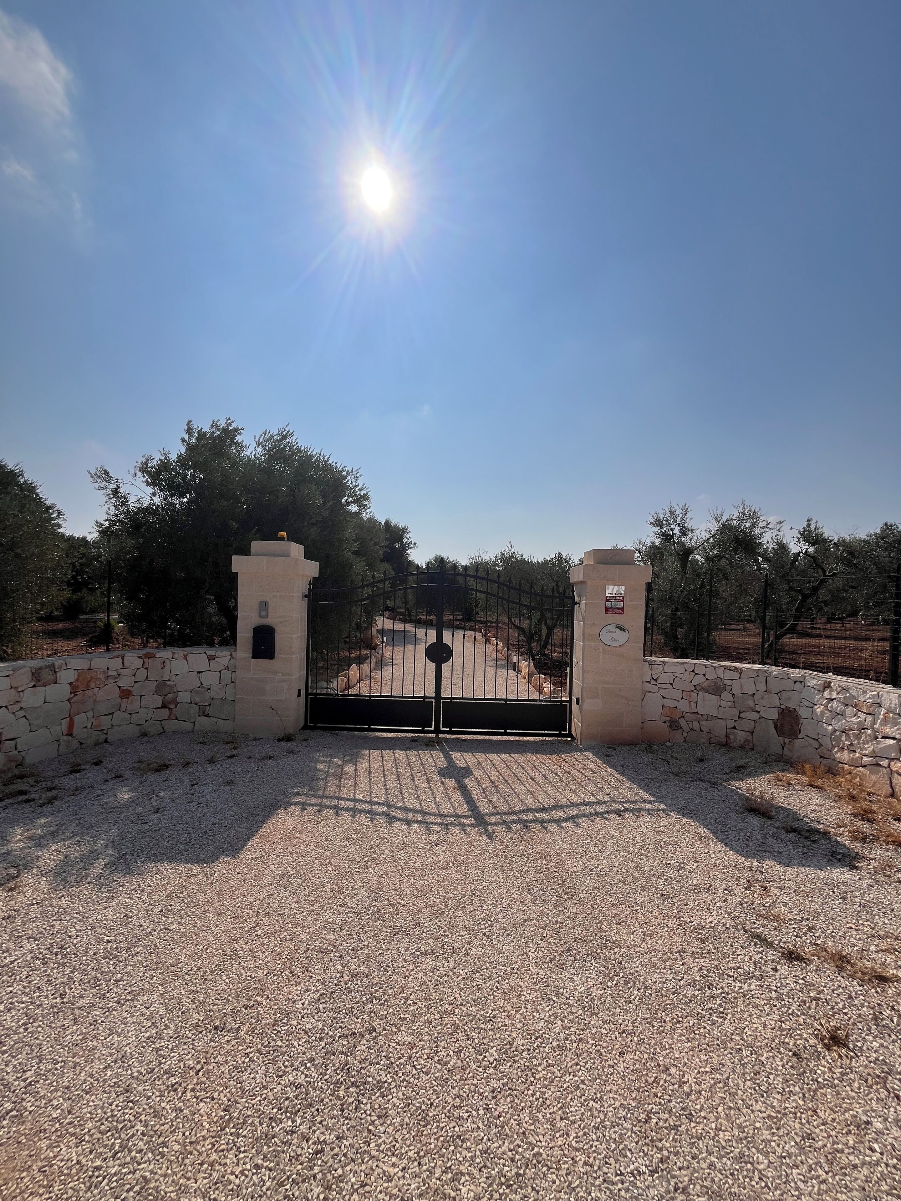 Una entrada protegida con un muro de piedra y una reja metálica, que da paso a un sendero rodeado de árboles.