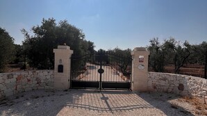 Une entrée fermée par un portail en métal et bordée d’un mur de pierre, qui donne sur un sentier entouré d’arbres.