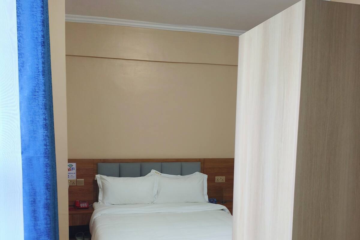 Una habitación de hotel con una cama, cabecera de madera, cortina azul y alfombra estampada.