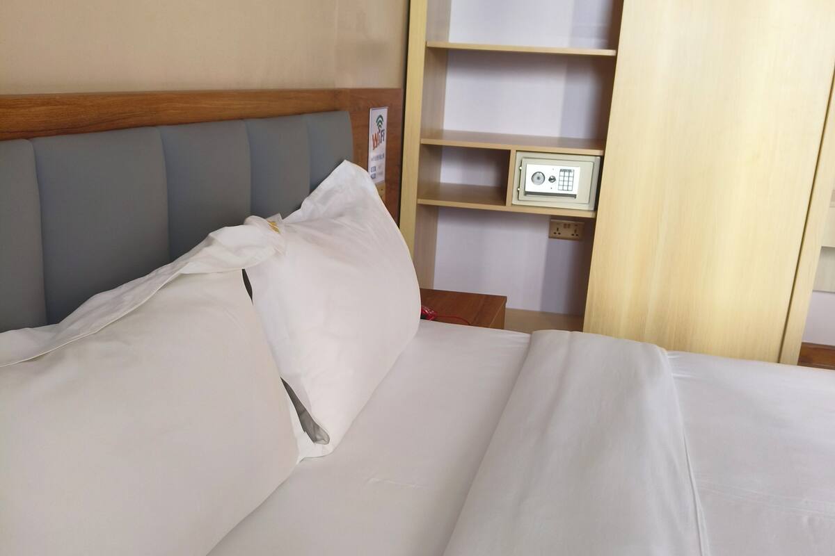 Una cama bien hecha con sábanas blancas, cabecera de madera y una estantería con un pequeño dispositivo electrónico encima.