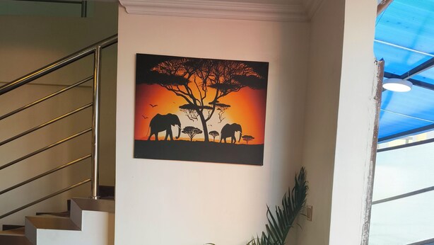 A staircase with a metal railing, a wall-mounted painting of elephants, a potted plant, a trash bin, and a table with water bottles and a tissue box.