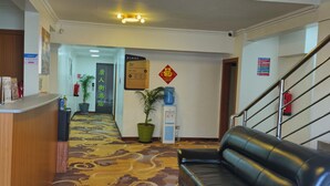 A hotel lobby with a reception desk, a black leather sofa, a patterned carpet, a staircase, and a wall-mounted sign.