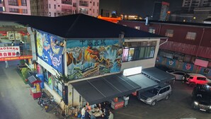 A building with a mural on the side, a covered outdoor seating area, and several parked cars.