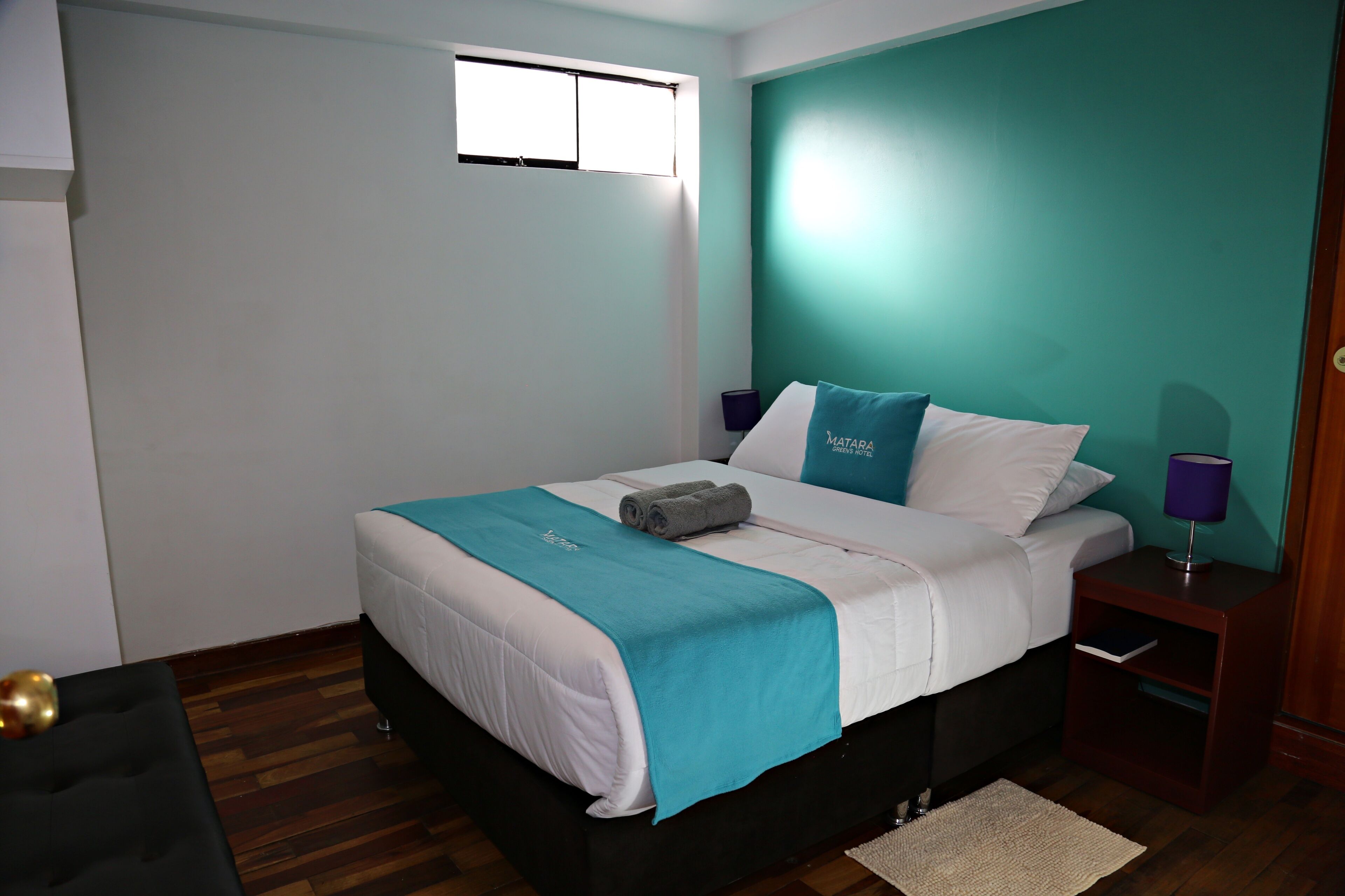 A neatly made bed with a teal and white bedspread, a small bedside table with a lamp, and a window above the bed.