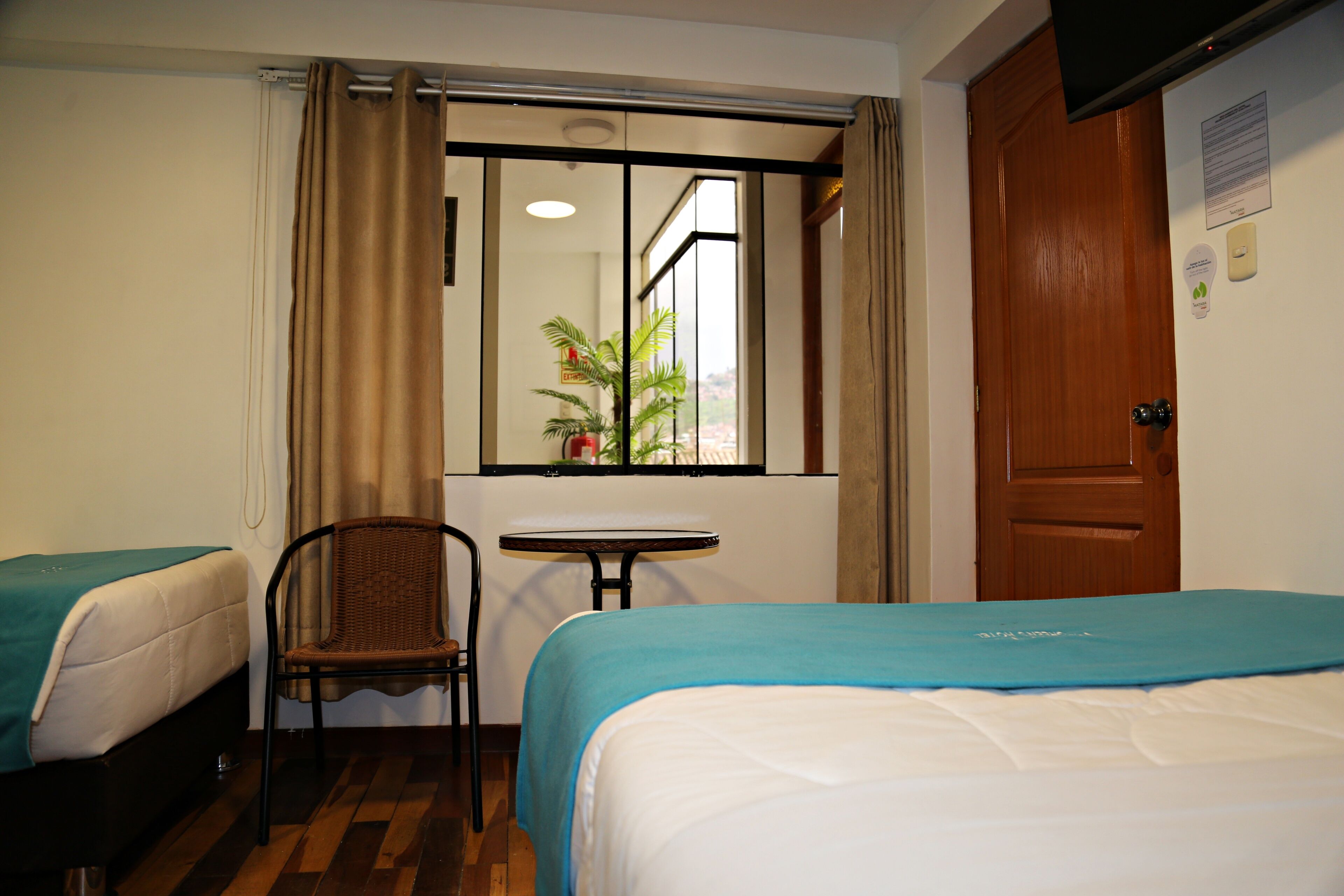 Habitación de hotel con dos camas, una mesita, una silla y un ventanal con cortinas.