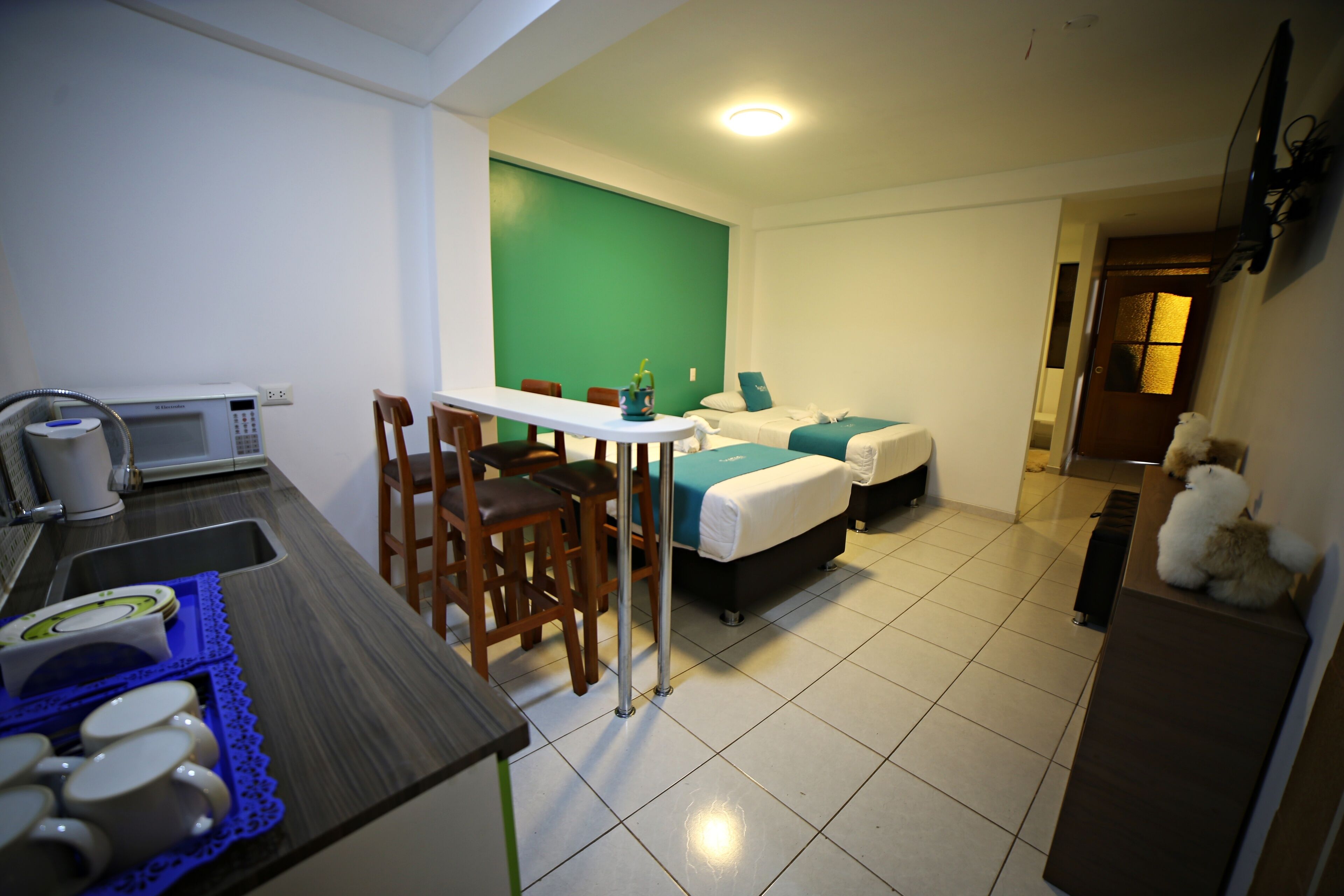 Une chambre d’hôtel compacte comprenant un coin cuisine, un espace repas et un lit avec un mur d’accent vert.