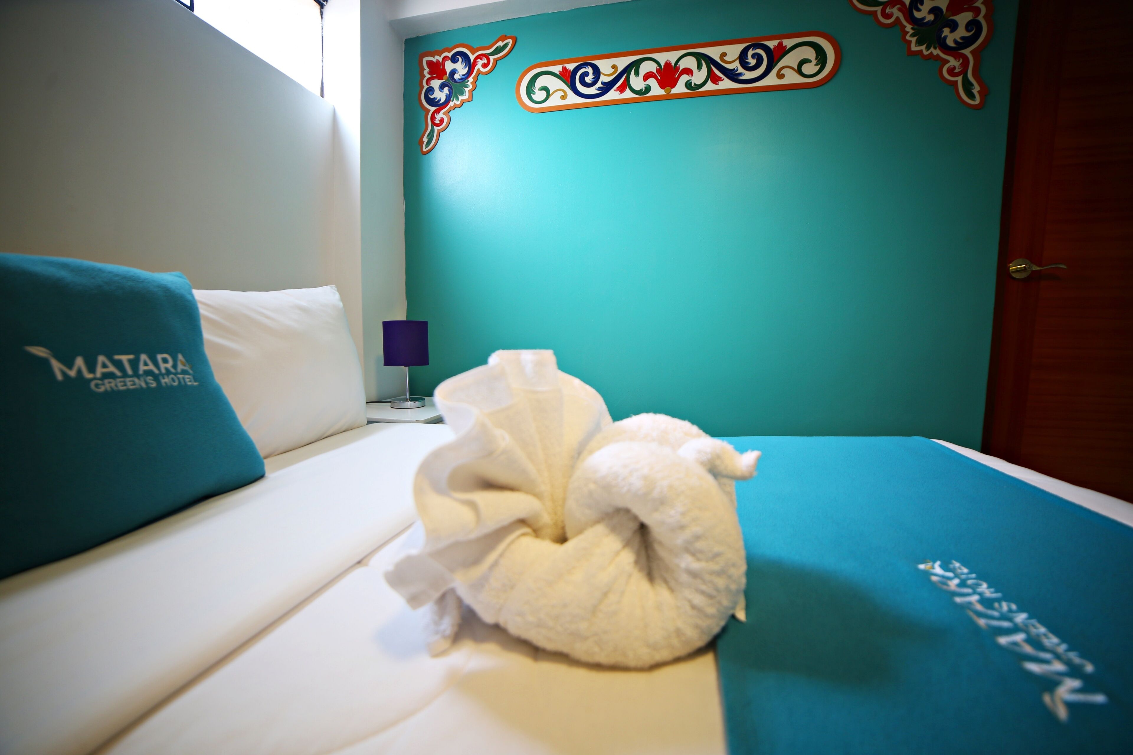 A hotel room with a teal accent wall, a neatly made bed with a white towel, and a pillow with the hotel name.