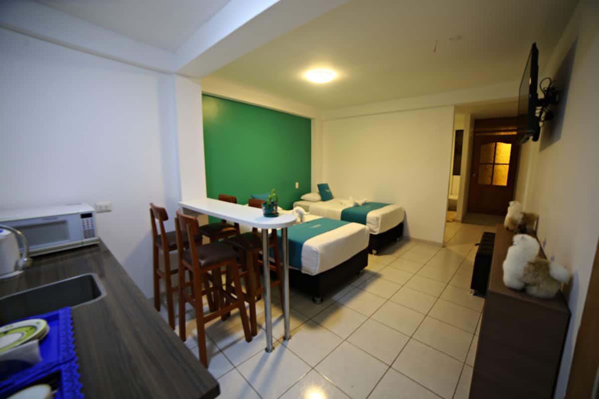 A compact hotel room with a kitchenette, a dining area, a bed, and a small seating area.