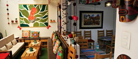 A cozy room with a sofa, wooden tables, and chairs, decorated with colorful pillows and wall hangings.