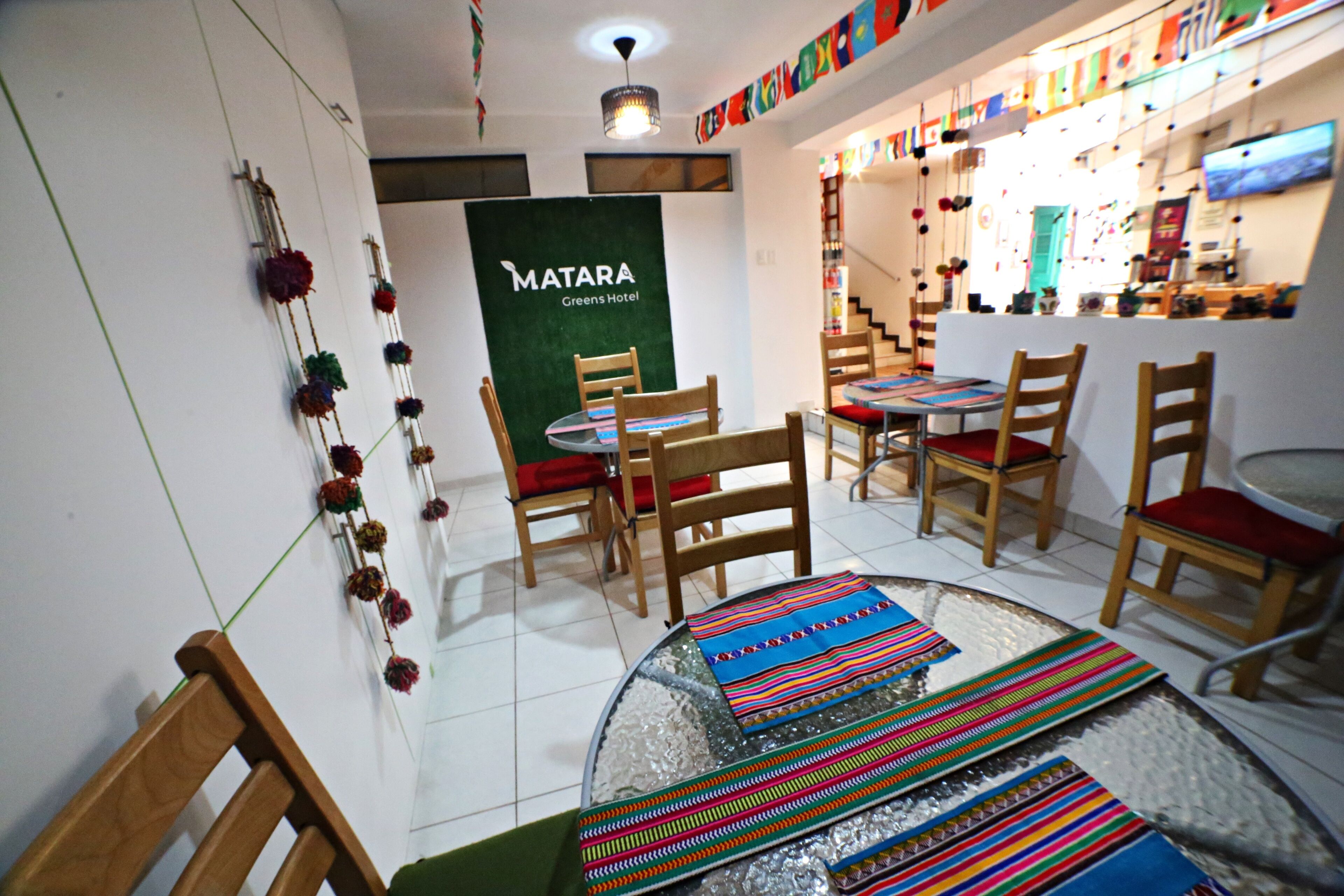 Habitación de hotel con una mesa redonda de vidrio, sillas de madera y una pared verde con el letrero 'MATARA Greens Hotel'.
