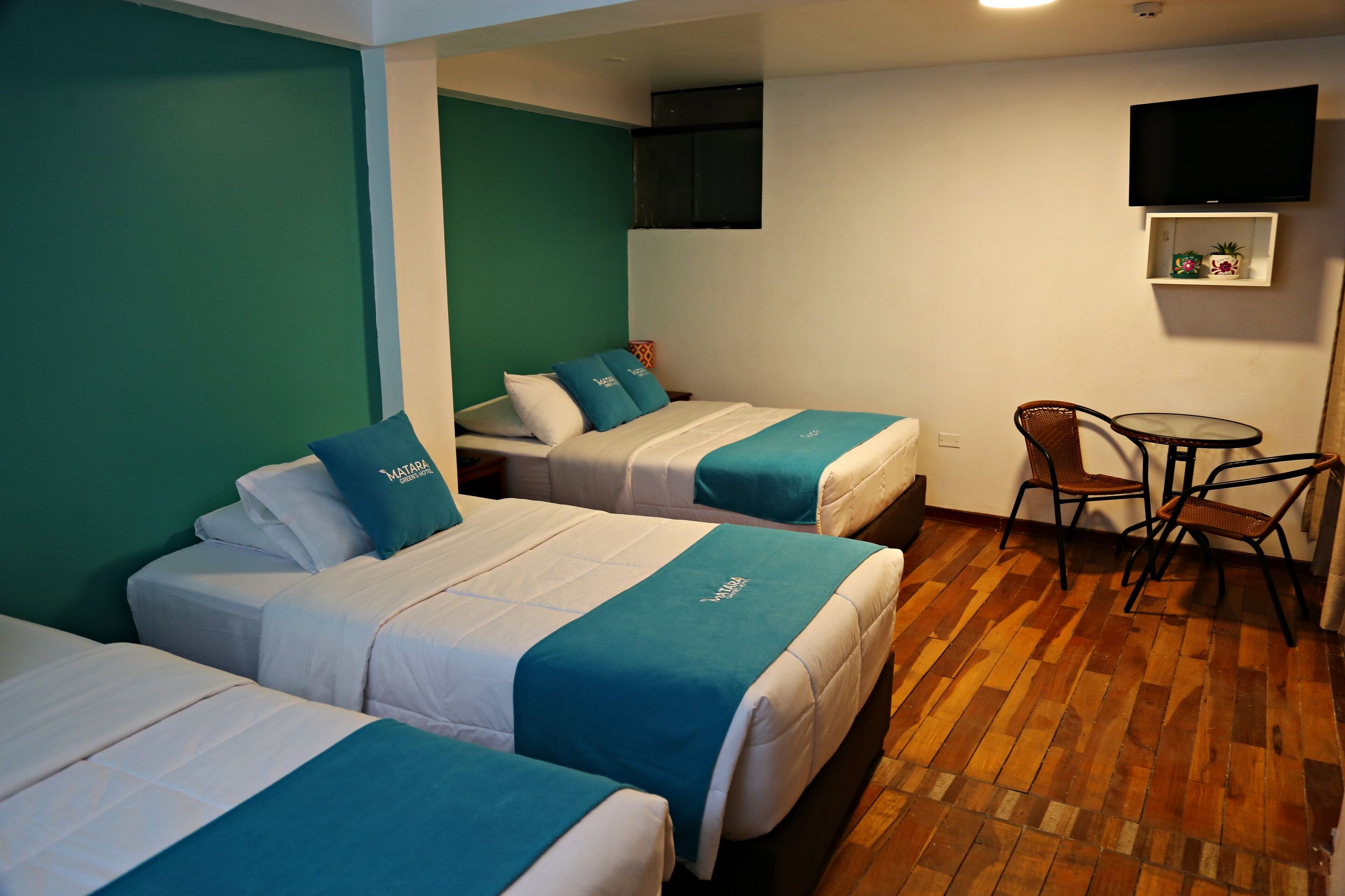 Ein Hotelzimmer mit zwei Betten, einem an der Wand montierten Fernseher, einem kleinen Tisch mit Stühlen und Holzboden.