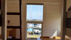 A room with a window overlooking a marina with boats. There is a wooden table and chairs, a small white appliance, and a shelf with a vase.