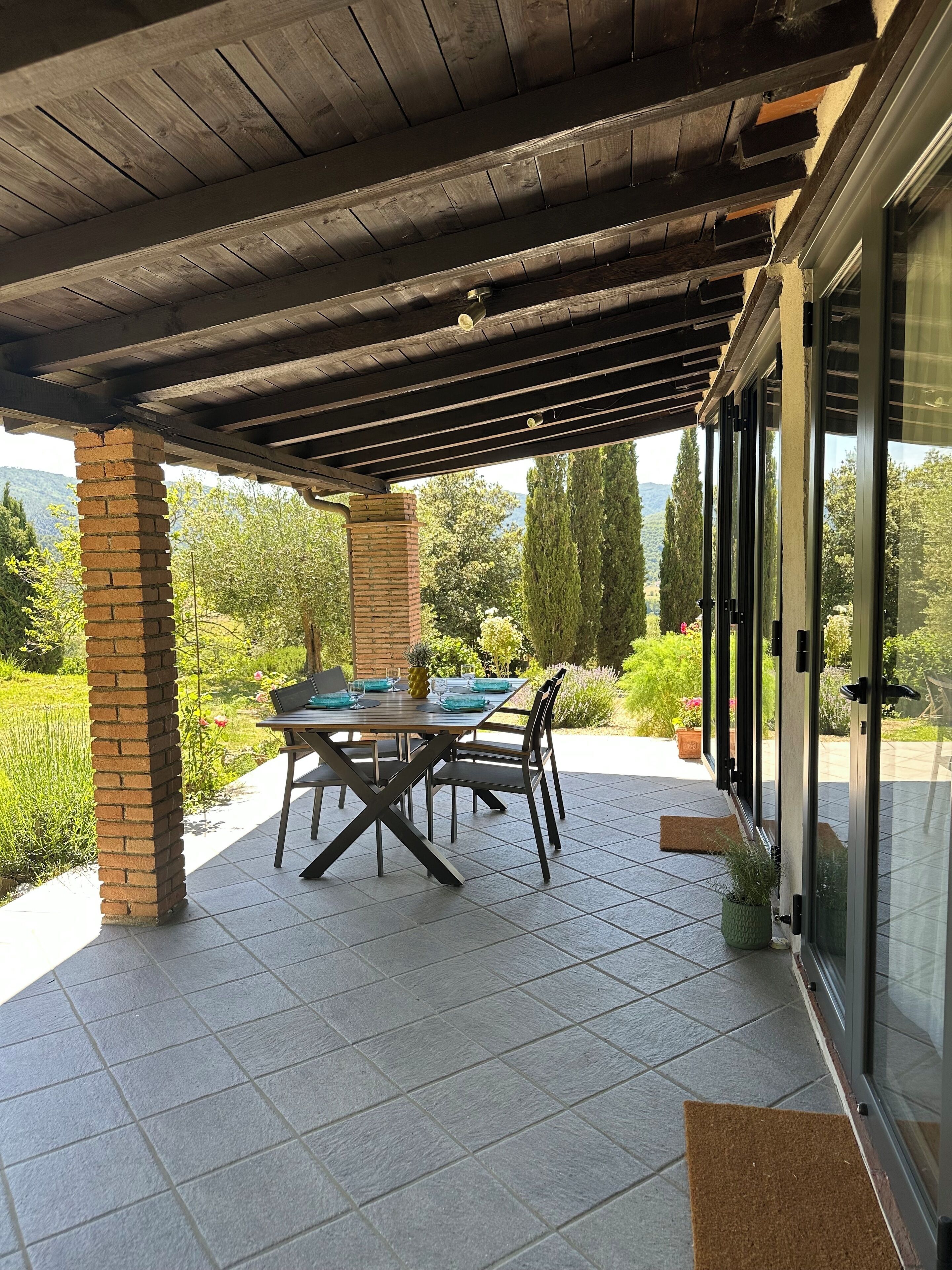 Eine überdachte Terrasse mit Tisch und Stühlen, Ziegelstützsäulen und Blick auf Grünflächen und Berge.