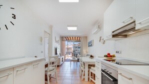 A narrow kitchen with white cabinets, a dining area with a table and chairs, and a view through an open door to the outside.