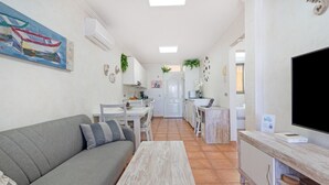A modern living room with a grey sofa, a wooden coffee table, and a TV on a wooden stand. There is a dining area with a white table and chairs, and a kitchen in the background.