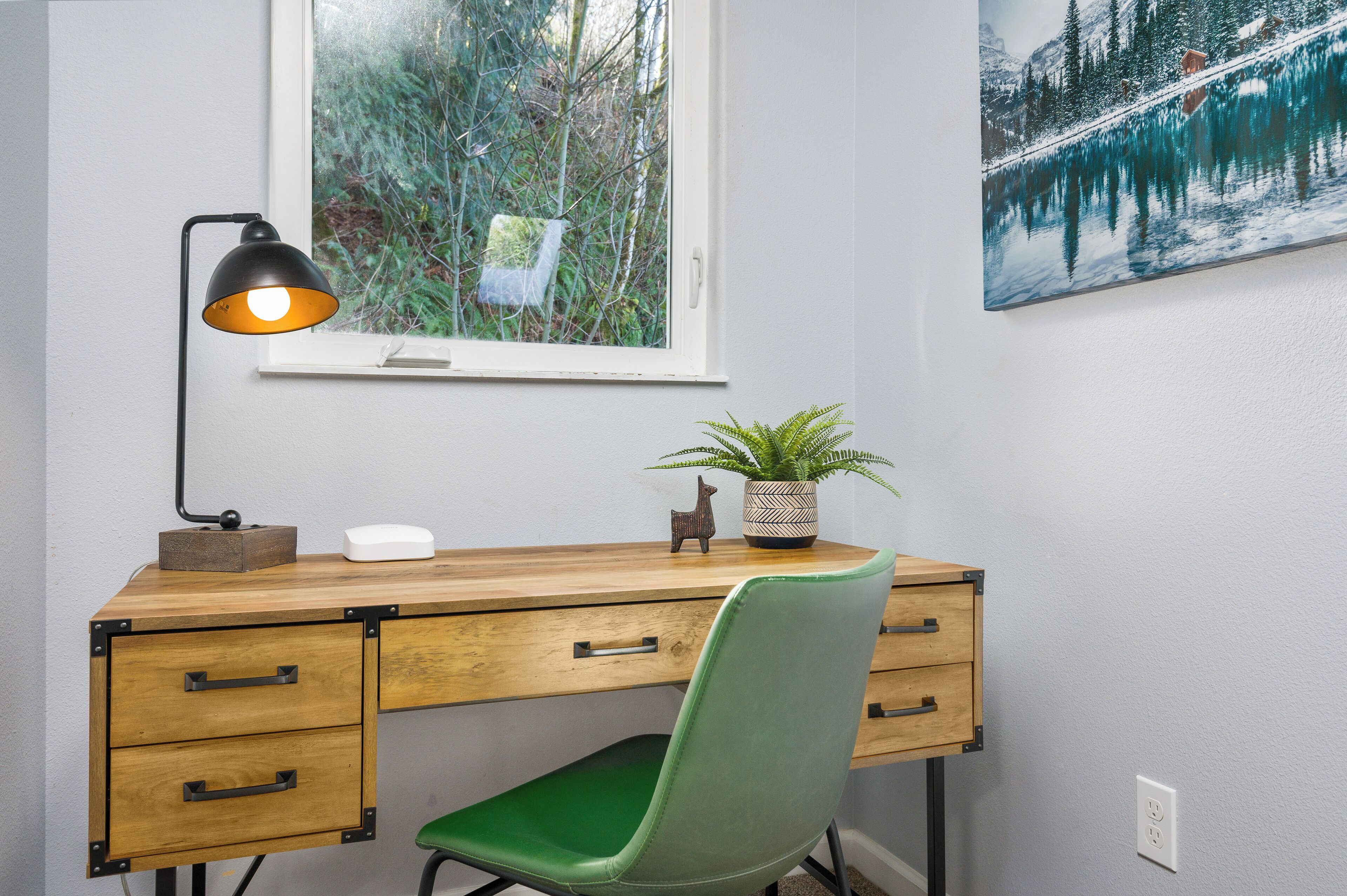 A wooden desk with drawers, a green chair, a desk lamp, a potted plant, and a window with a view of trees.