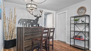A dining area with a wooden table and chairs, a decorative deer wall art, a chandelier, and a bookshelf with books and a plant. - Dog Friendly! Hot Tub~Game Room~Fire Pit~3BR, 2BA~High Speed WIFI~8 Guests Max (Mount Hood Village)