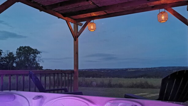 A hot tub with a waterfall feature, under a wooden pergola with hanging lanterns, overlooking a grassy landscape.