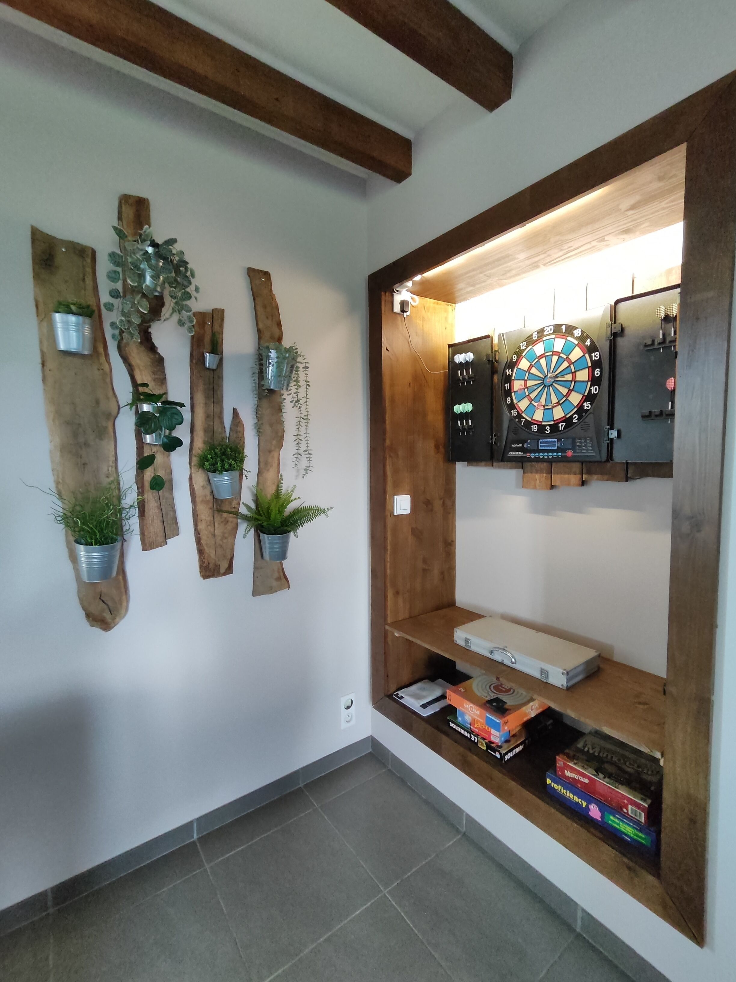A room with a dartboard, wooden wall decor with plants, and a shelf with books and electronic devices.