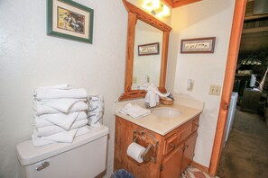 A bathroom with a toilet, sink, and mirror. There are towels stacked on the toilet, a basket on the sink, and a framed picture on the wall. - Cardinal Ridge (Big Bear City)