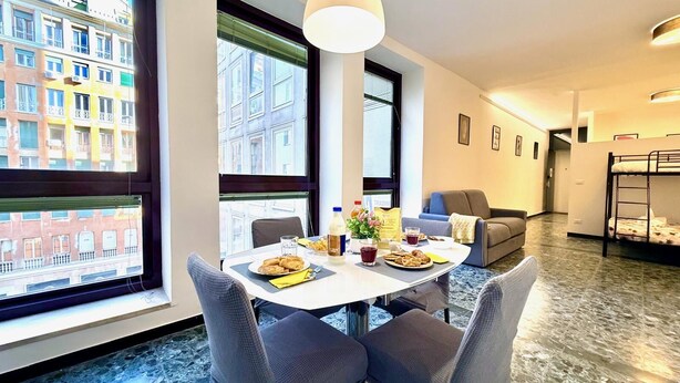 Ein modernes Hotelzimmer mit Essbereich, einem Tisch für zwei Personen, einem großen Fenster mit Stadtblick, einem Stockbett und einer gemütlichen Sitzecke.