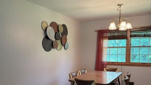 Un comedor con una mesa redonda, seis sillas, un tapete estampado, un adorno en la pared y una ventana con cortinas.