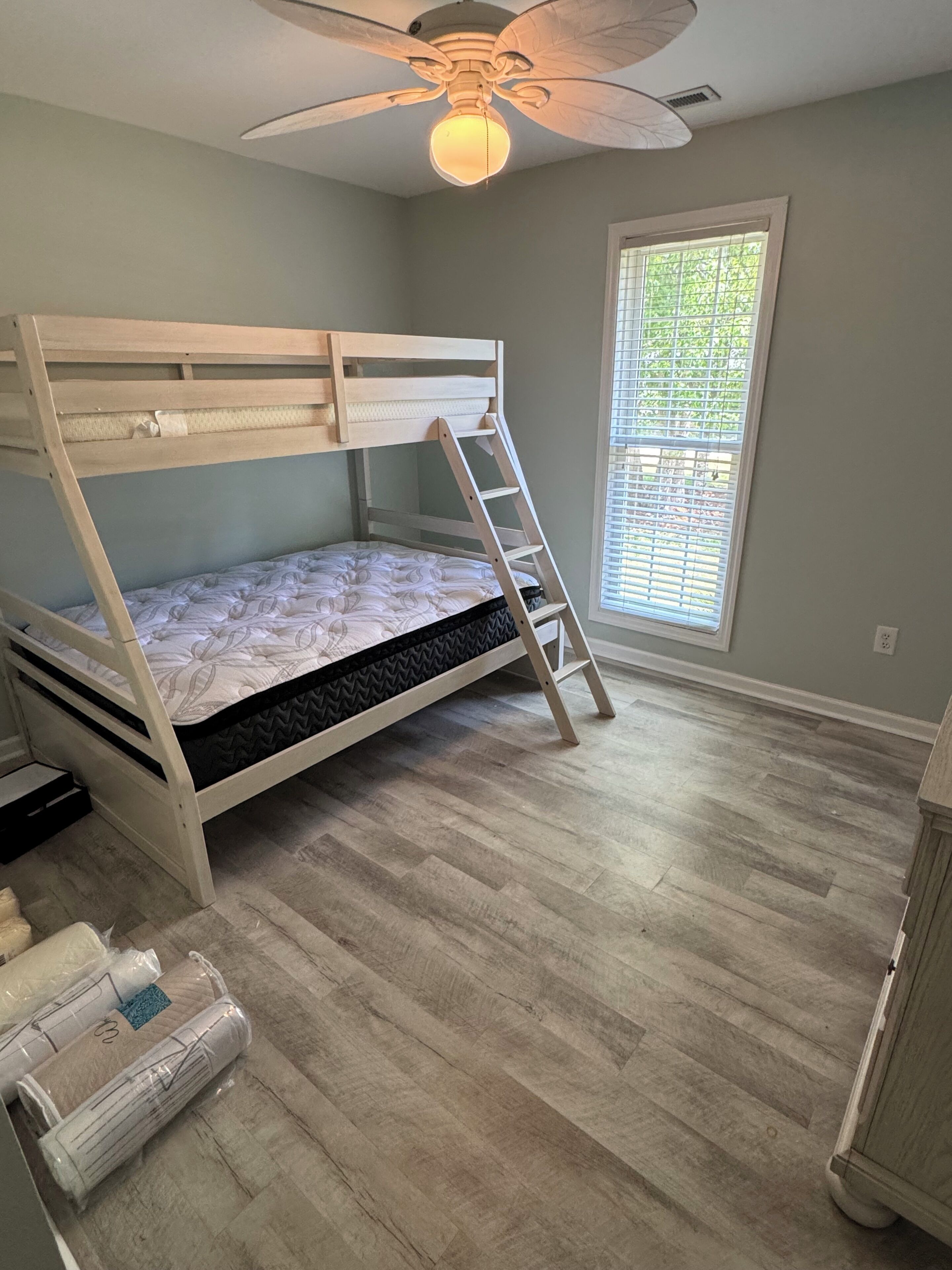 A bunk bed room with a ceiling fan, a window with blinds, and a wooden floor.