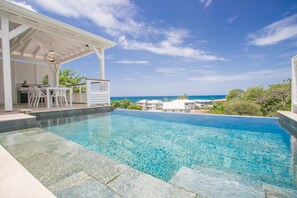 Piscina infinita de color azul claro con un pérgola blanca y zona de estar exterior con vistas al océano.