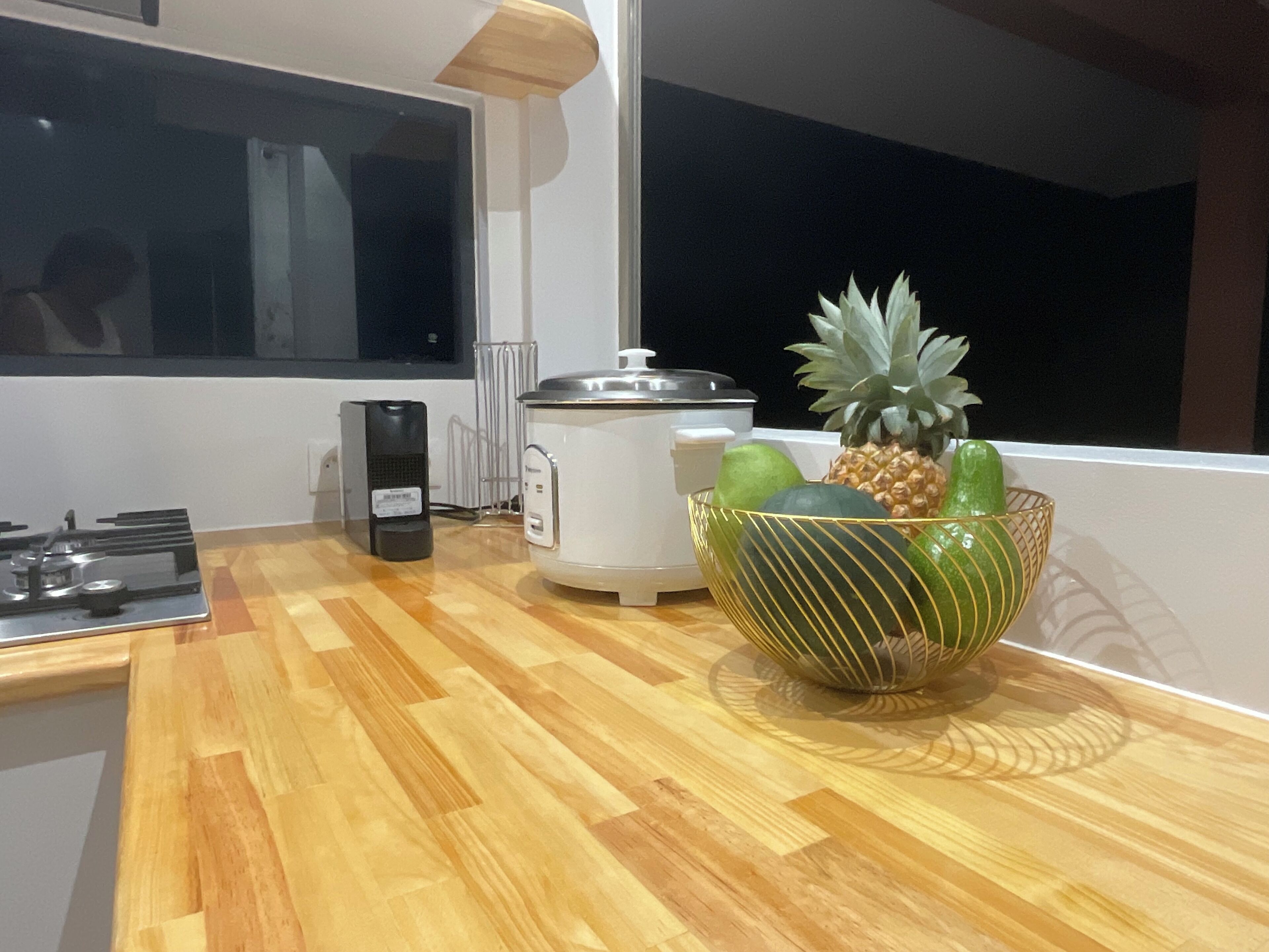 Une cuisine avec un plan de travail en bois, une machine à café, un cuiseur à riz et un panier à fruits.