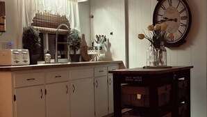 Een keuken met witte kastjes, een vintage klok en een tafel met een bord waarop staat 'Save Water Drink Beer'.