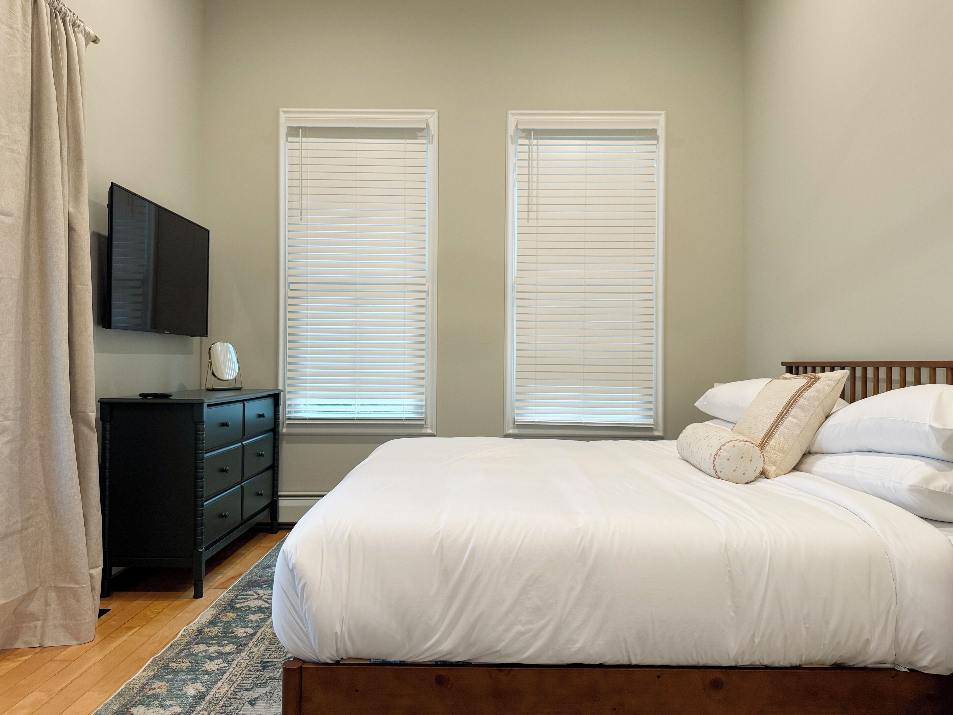 A bedroom with a bed, a dresser, a TV, and two windows with blinds.