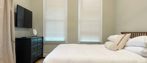 A bedroom with a bed, a dresser, a TV, and two windows with blinds.
