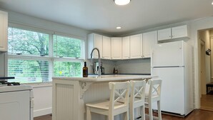 A kitchen with white cabinets, a white refrigerator, a bar area with white chairs, and a window with blinds. - Historical Charm-Saugatuck City-Sleeps 8 (Saugatuck)