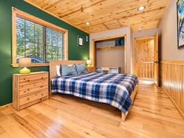 A bedroom with a wooden ceiling, a bed with a plaid bedspread, a nightstand with a lamp, a dresser, and a window with blinds.