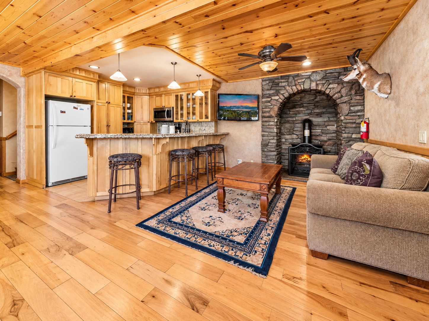 Un interior de madera con cocina, taburetes de bar, chimenea de piedra y sofá.