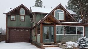 En tvåvånings timmerstuga med ett garage, stora fönster och ett snötäckt landskap.