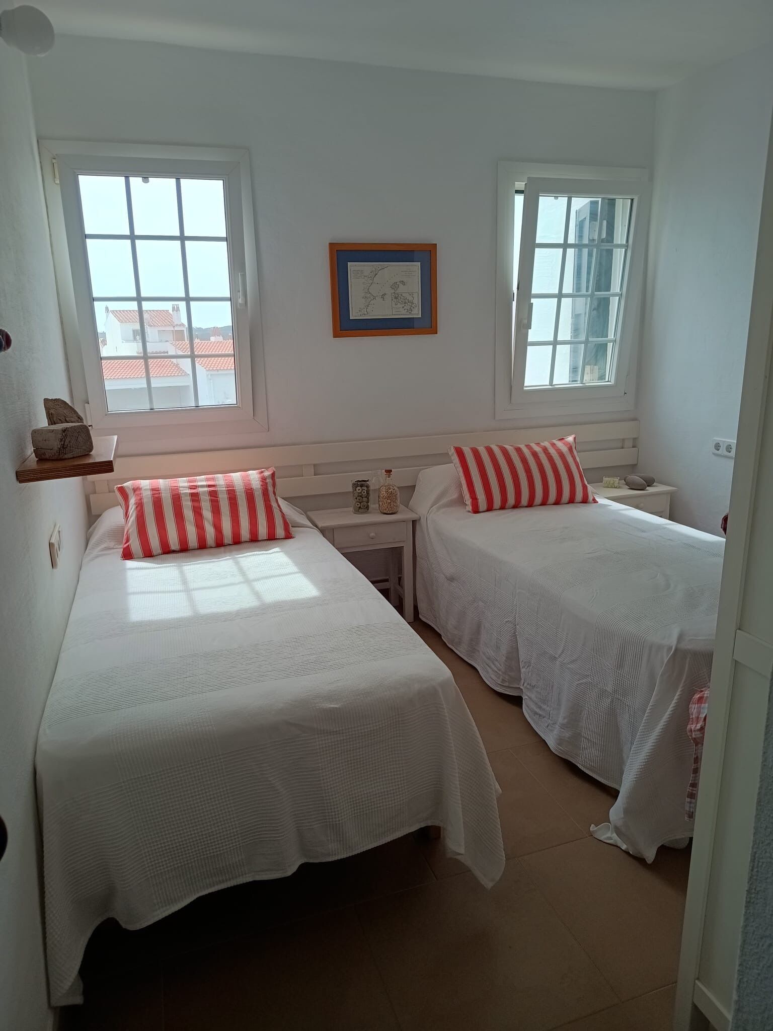 A room with two single beds, a small table, a shelf with decorative items, and a framed map on the wall.